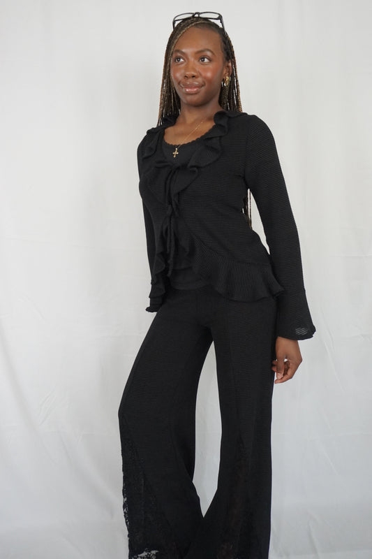 A woman modeling a black two-piece outfit consisting of a long-sleeve ruffled blouse and matching wide-leg pants, standing against a white studio backdrop.